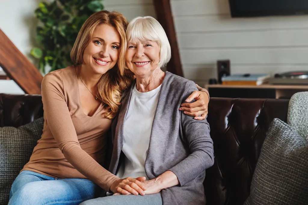 Portrait of senior aged mother and middle aged daughter smiling together on the couch at home